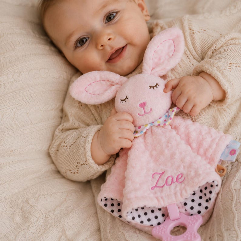 May include: A soft pink plush security blanket featuring a rabbit head with embroidered closed eyes and a nose. The blanket has a multi-colored polka dot neck ruffle, a black and white polka dot underside, and a pink star-shaped teether attached.