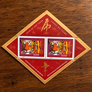 Könnte beinhalten: Eine Gedenkbriefmarkenbogen mit zwei Briefmarken mit einer Tigerillustration. Die Briefmarken sind vor einem lila Hintergrund. Das Blatt ist rot mit goldenen Akzenten und Text in Englisch und Französisch, zum Jahr des Tigers.