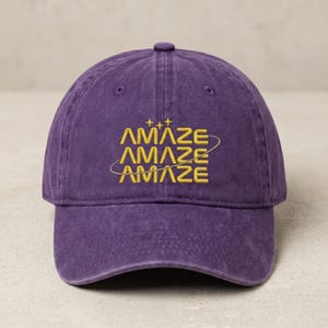 May include: A purple baseball cap with the word "AMAZE" embroidered in yellow. The word is repeated three times, with stars and a curved line above the top word. The cap has a curved brim and a button on top.