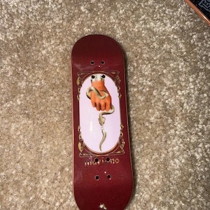 Puede incluir: Una tabla de dedos roja con un gráfico de una mano naranja sosteniendo una serpiente, dentro de un marco decorativo. La tabla de dedos tiene la palabra "mariano" impresa. El fondo es de color rosa claro.