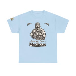 Könnte beinhalten: Hellblaues T-Shirt mit einer detaillierten Illustration einer historischen Figur und dem Text "trust me, I'm a - Medicus". Die Grafik ist in Braun- und Beigetönen gehalten. Das T-Shirt hat einen Rundhalsausschnitt.