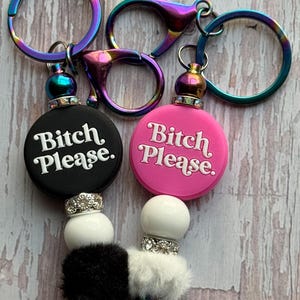 May include: Two keychains with iridescent hardware. One keychain is black with the text "Bitch Please." The other is pink with the same text. Both have white beads, a fluffy accent, and a metallic bead.