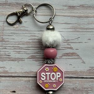 May include: A keychain featuring a silver lobster clasp and ring, a white faux fur pom-pom, a pink bead, a silver rhinestone spacer, and a pink octagonal pendant. The pendant displays the white text STOP OVERTHINKING IT with yellow smiley faces and white flower accents.