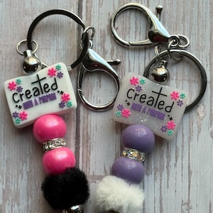 May include: Two keychains with the text "Created with a Purpose". One keychain has pink beads and a black pom-pom, the other has purple beads and a white pom-pom. Both have silver hardware.