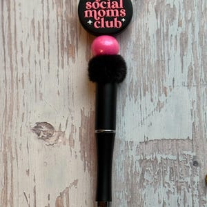 May include: A black pen with a decorative top that reads "anti social moms club" in pink lettering. The pen has a pink bead, a black fluffy accent, and a silver tip. The pen is on a light wood grain surface.