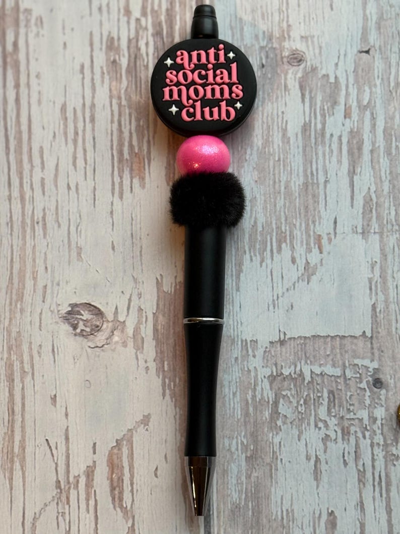 May include: A black pen with a decorative top that reads "anti social moms club" in pink lettering. The pen has a pink bead, a black fluffy accent, and a silver tip. The pen is on a light wood grain surface.