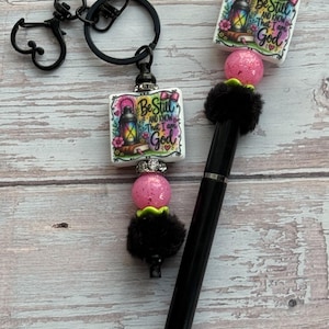 May include: A black ink pen and a matching keychain, both featuring a square bead with a lantern illustration and the text "Be Still AND KNOW That I am God." Each item includes a pink glitter bead, a black fuzzy bead, and a silver accent bead.