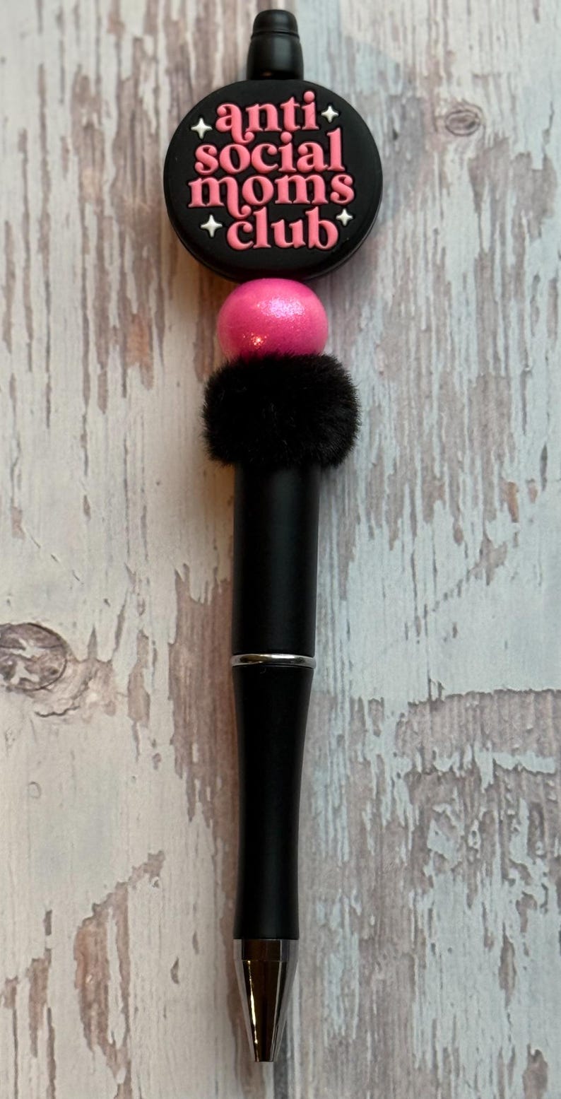 May include: A black pen with a decorative top that reads "anti social moms club" in pink lettering. The pen has a pink bead, a black fluffy accent, and a silver band.