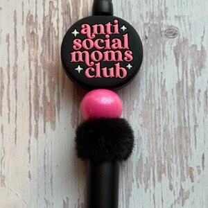 May include: A black pen with a decorative top that reads "anti social moms club" in pink lettering. The pen has a pink ball and a black fluffy accent.