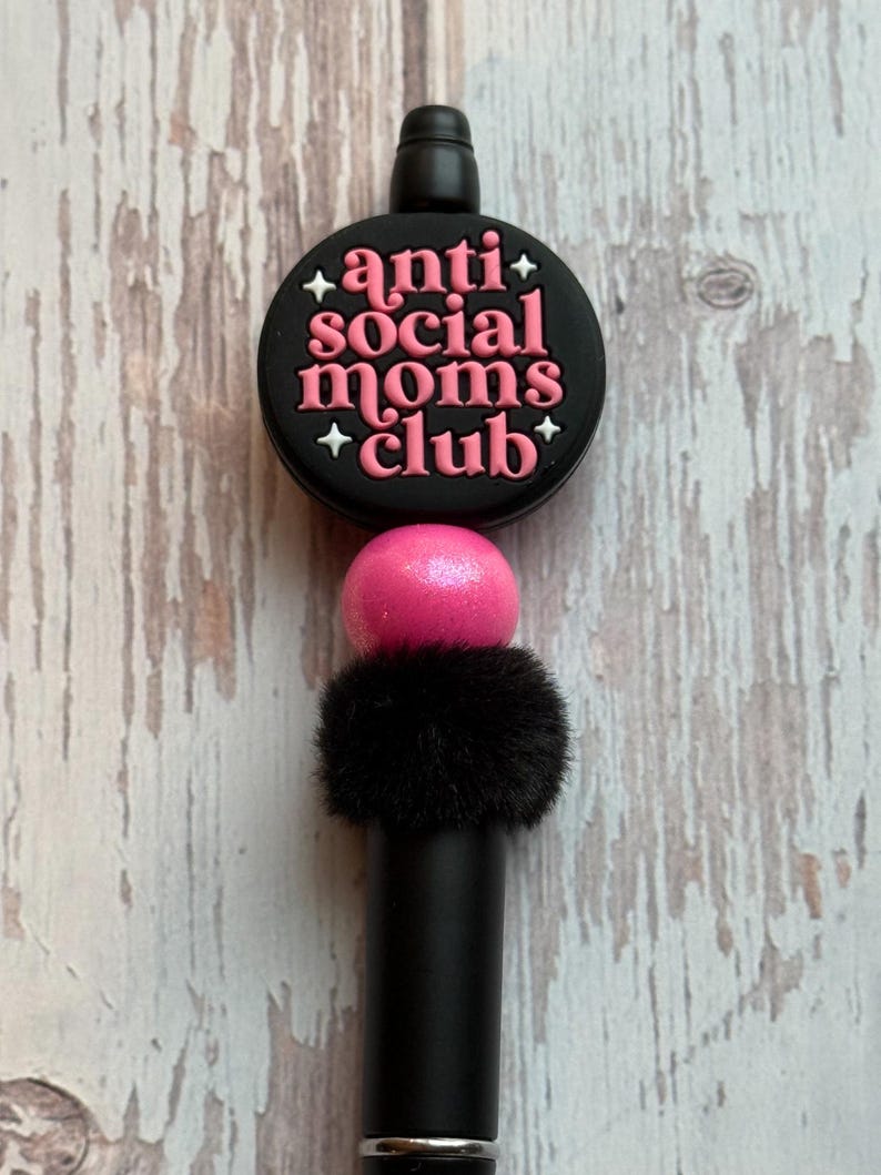 May include: A black pen with a decorative top that reads "anti social moms club" in pink lettering. The pen has a pink ball and a black fluffy accent.