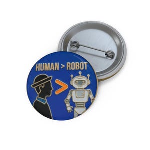 May include: A round button pin with the text "HUMAN > ROBOT" in white. The pin features a silhouette of a person in a hat and a robot on a blue background. The pin is silver and has a pin back.