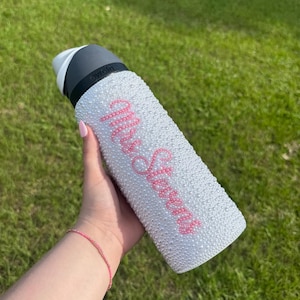 Puede incluir: Una botella de agua blanca cubierta de pequeñas decoraciones blancas con forma de perlas. La botella tiene una tapa negra y gris y presenta el texto rosa "Mrs. Stevens". La botella se sostiene sobre un fondo de hierba verde.