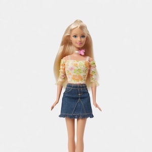 Peut inclure: Poupée blonde aux yeux bleus, vêtue d'un haut fleuri à épaules dénudées et d'une jupe en jean à ourlet effiloché. Elle porte un collier ras du cou rose et un accessoire floral rose. Fond blanc.