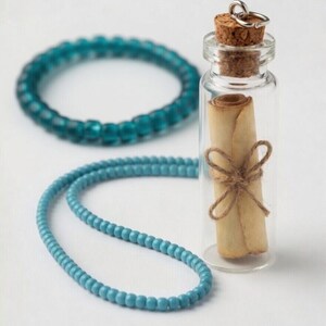 May include: A clear glass bottle with a cork stopper holds a rolled-up scroll tied with twine. A turquoise beaded bracelet and necklace are in the background. The items are set against a white backdrop.