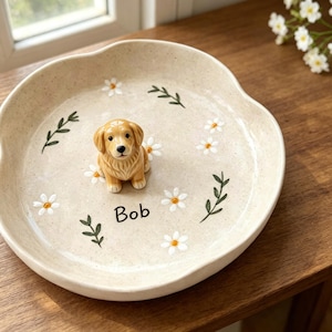 May include: A beige ceramic dog bowl with a raised rim, featuring a small golden retriever figurine in the centre. The bowl is decorated with white daisies and green leaf accents. The name "Bob" is written on the bowl.