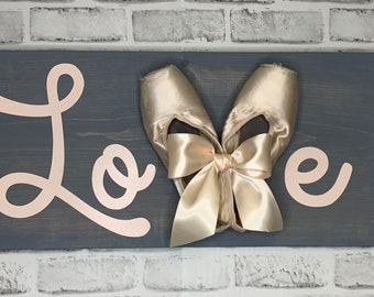 Peut inclure: Une pancarte en bois avec le mot "Love" écrit en lettres roses. Deux chaussons de ballet sont liés ensemble avec un grand nœud en satin au centre de la pancarte.