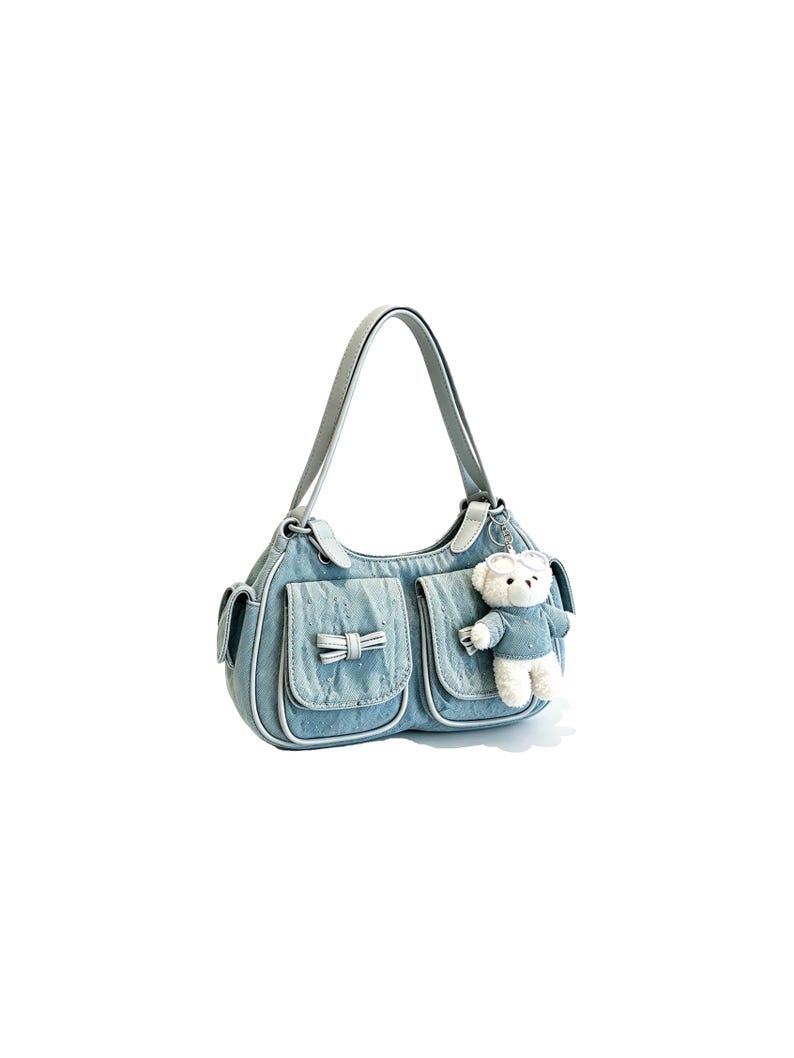 Puede incluir: Bolso de hombro de mezclilla azul claro con dos bolsillos delanteros y un peque&ntilde;o detalle de lazo. El bolso tiene un ribete blanco y un asa a juego. Un peque&ntilde;o llavero de oso de peluche cuelga del lateral.