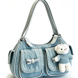 Puede incluir: Un bolso de mano de mezclilla azul claro con asa y ribete grises. El bolso presenta dos bolsillos delanteros con lazos y un peque&ntilde;o llavero de oso de peluche blanco con gafas y un atuendo de mezclilla a juego.