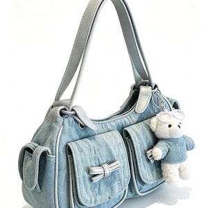 Puede incluir: Bolso de mano de mezclilla azul claro con detalles plateados y un lazo blanco en el bolsillo delantero. Cuenta con un asa gris claro y un peque&ntilde;o colgante de oso de peluche con gafas y un su&eacute;ter de mezclilla.
