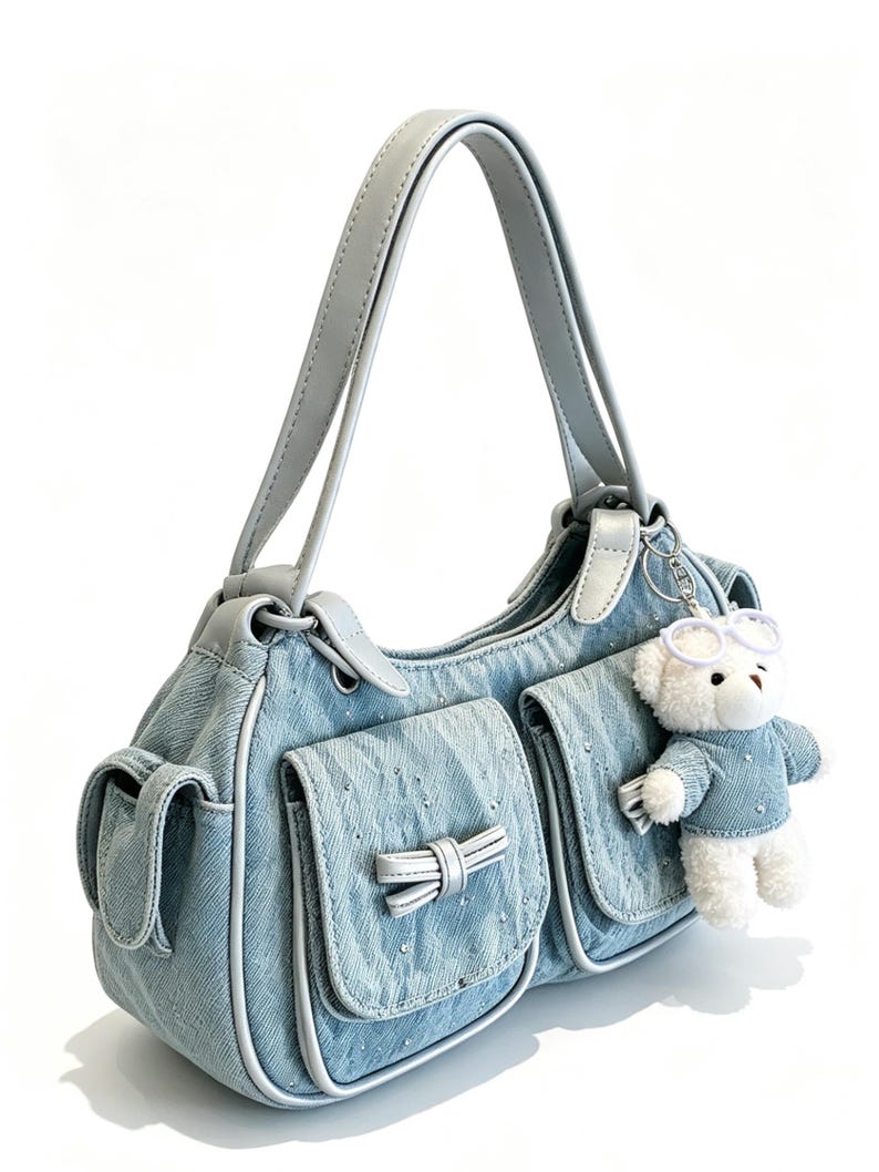 Puede incluir: Bolso de mano de mezclilla azul claro con detalles plateados y un lazo blanco en el bolsillo delantero. Cuenta con un asa gris claro y un peque&ntilde;o colgante de oso de peluche con gafas y un su&eacute;ter de mezclilla.