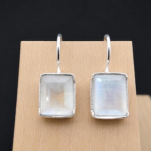 Peut inclure: Une paire de boucles d'oreilles rectangulaires avec un cadre argenté et une pierre claire et irisée. Les boucles d'oreilles sont présentées sur un présentoir en bois sur fond noir. Les pierres ont un éclat nacré subtil.