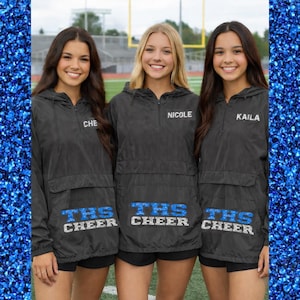 May include: Three smiling individuals wearing black hooded jackets with the text "THS CHEER" in blue and white. Each jacket has a name embroidered on the chest: "CHE", "NICOLE", and "KAILA". The background shows a football field.