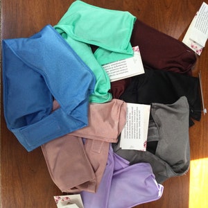 May include: A collection of colorful, fabric-covered eye masks in shades of blue, green, purple, pink, gray, black, and burgundy. Each mask has a rectangular shape with a curved top and a strap. Some masks have attached tags.