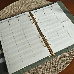 May include: Open planner with a weekly schedule layout. The pages are white with gray lines and dots, and the planner is held open by gold rings. The cover is a dark green leather-like material, and a light blue pen is visible on the left.