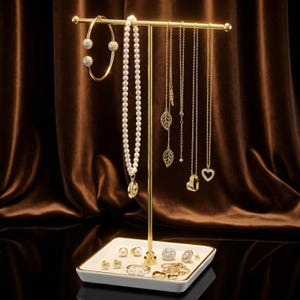 May include: A gold-colored jewelry display stand holding various necklaces, a bracelet, earrings, and rings. The necklaces include a pearl strand and delicate chains with leaf and heart pendants. The base is a white tray with gold trim.