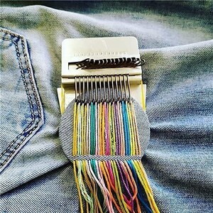 May include: A white and metal weaving tool with colorful threads in a variety of colors, including yellow, blue, green, and purple. The tool is set on a denim background. The threads are hanging down from the tool.