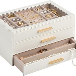 May include: A white jewelry box with a glass top lid, two pull-out drawers, and gold-toned rectangular handles. The interior is lined with beige velvet and contains various compartments for rings, necklaces, watches, and bracelets.