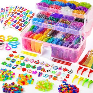 May include: A multi-tiered clear plastic storage case filled with assorted colorful rubber bands, accompanied by sheets of number and letter stickers, various shaped charms, plastic beads, small hooks, and colorful tassels for crafting projects.