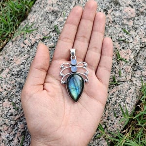 May include: A silver pendant with a teardrop-shaped labradorite stone, displaying iridescent blue and green hues. Two smaller, round, light blue stones and decorative silver accents complete the design. The pendant is held in an open hand against a natural background.