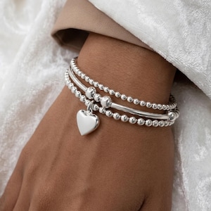 Puede incluir: Tres pulseras plateadas apiladas en una muñeca. Una pulsera tiene un dije de corazón, las otras tienen un diseño de cuentas. Las pulseras están hechas de metal plateado brillante y se exhiben sobre un fondo blanco y beige.
