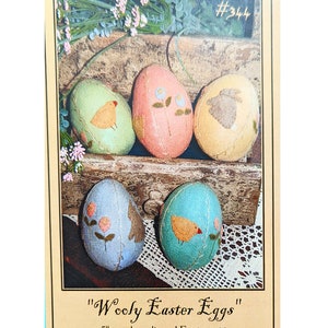 May include: A framed image showing six colourful, wool appliquéd Easter eggs, each with unique designs of birds, flowers, and a bunny. The eggs are displayed on a rustic wooden shelf. The text reads "Wooly Easter Eggs" and "Country Stitches". Each egg is approximately 13 cm.