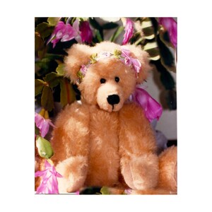 May include: A light brown plush teddy bear with a fuzzy texture, featuring a black stitched nose and eyes. The bear is adorned with a decorative headband made of small pink and green fabric flowers and ribbons tied around its head.