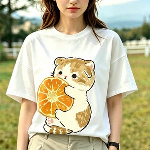 May include: White t-shirt featuring a cartoon cat holding a large orange slice. The cat has brown and white fur, and the orange is a bright orange color. The shirt has short sleeves and a relaxed fit.