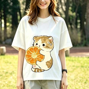 May include: White t-shirt featuring a cartoon cat holding an orange slice. The cat has brown and white fur, and the orange is a bright orange color. The shirt is a casual, oversized style.