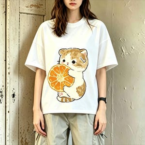 May include: White t-shirt featuring a cartoon cat holding a slice of orange. The cat has brown and white fur. The t-shirt is a casual, short-sleeved style.