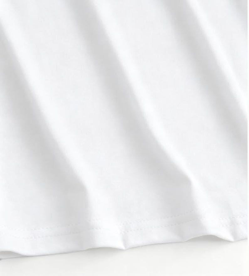 May include: Close-up of a white t-shirt, showing the fabric's texture and the stitched hemline. The fabric has subtle diagonal folds, creating shadows and highlights. The image focuses on the clean, crisp appearance of the garment.