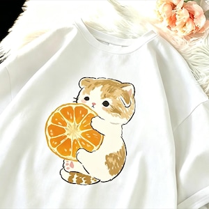 May include: White t-shirt featuring a cartoon cat holding a large orange slice. The cat has brown and white fur, with a cute expression. The orange slice is bright orange with visible segments. The shirt is made of a soft material.