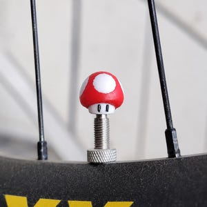 Puede incluir: Un tapón de válvula con forma de seta roja y blanca con un vástago de metal plateado roscado. La parte superior de la seta presenta manchas circulares blancas sobre un fondo rojo, y la base es blanca con dos pequeños ojos negros.