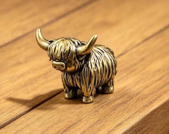 May include: A small, solid brass figurine of a Highland cow. The metal sculpture features a textured, shaggy coat, curved horns, and a detailed face with a prominent snout. The finish is a polished, warm metallic gold with dark patina.