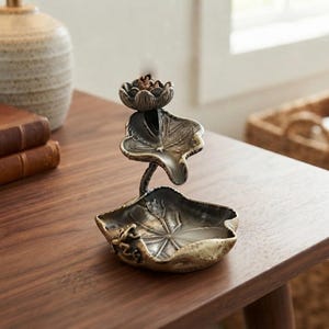 Pode incluir: Um queimador de incenso metálico com uma flor de lótus esculpida no topo, uma secção central curva em forma de folha e uma base larga e rasa em forma de nenúfar. O acabamento é num tom de bronze antigo escuro com reflexos claros.