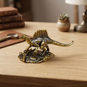Puede incluir: Una figura de dinosaurio Spinosaurus de metal en tono dorado sobre una base texturizada. La escultura presenta una vela dorsal prominente, una cola larga y una textura de piel detallada, capturando a la criatura en una pose dinámica.
