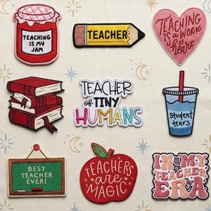 May include: A collection of colorful embroidered patches with teacher-themed designs. Patches include a pencil, books, a heart, a jar of jam, an apple, and phrases like "Teaching is my Jam" and "Best Teacher Ever!" on a cream-colored fabric.