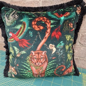 May include: Square cushion with a dark green jungle design featuring a cougar, parrots, and monkeys. The cushion has black fringe trim around the edges. The design includes various jungle animals.