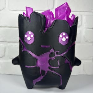 May include: A black planter shaped like a stylized creature with a matte finish. The top features translucent purple crystal-like protrusions. The front displays a cutout of a purple figure with four-dot eyes and a small horizontal mouth.