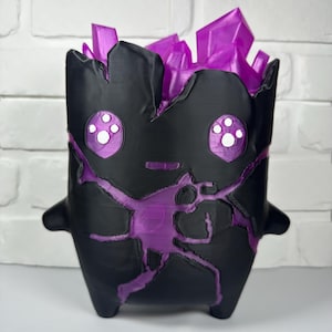 Puede incluir: Una maceta negra con acabado mate que presenta un diseño recortado de un personaje en una pose dinámica. El interior y las estructuras sobresalientes en forma de cristal en la parte superior son de color morado translúcido. El personaje tiene ojos grandes.
