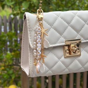 Puede incluir: Un bolso blanco acolchado con cadena y cierre dorados. Un dije dorado con la palabra "REJOICE" en cuentas blancas cuelga de la cadena, junto con estrellas de mar, conchas y perlas.